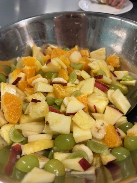 A bowl of mixed fruit salad contains apples, oranges, grapes, and bananas. The bowl is stainless steel.