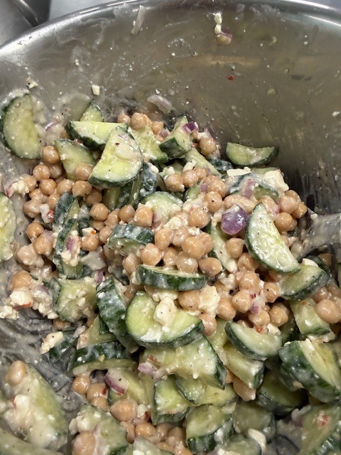 A bowl contains a mix of chopped cucumbers, chickpeas, onions, and possibly other vegetables, with a creamy dressing.