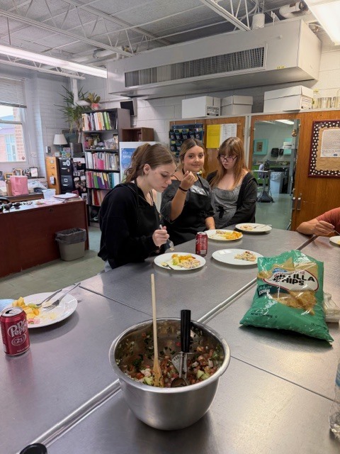 A group of people sit at a table eating food, with a bowl of ingredients in the center.