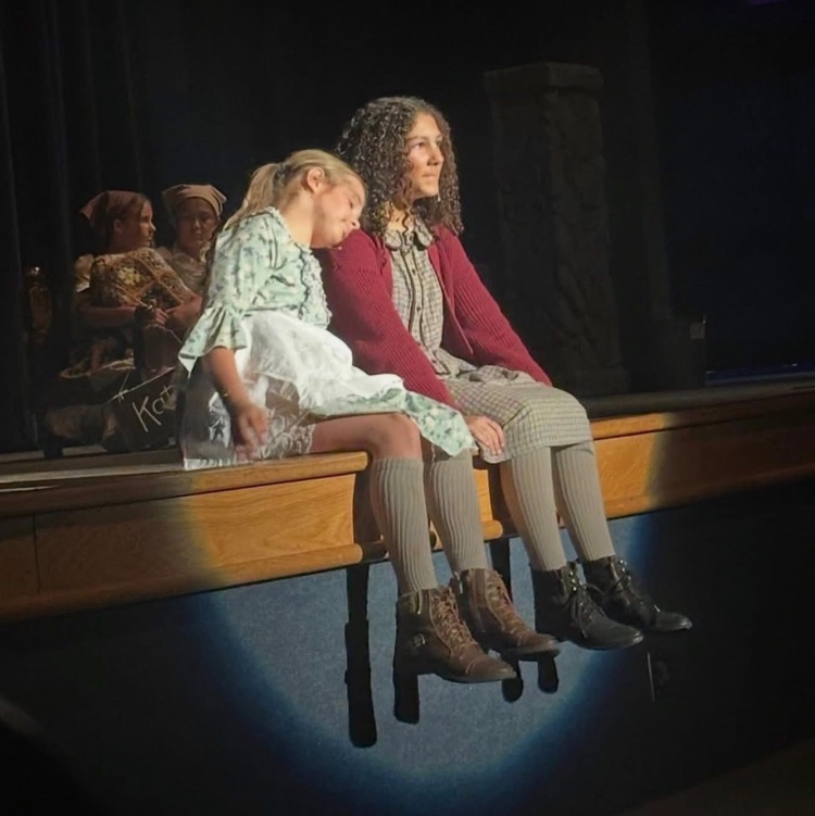 Two girls dressed as orphans sit on edge of stage in spotlight  