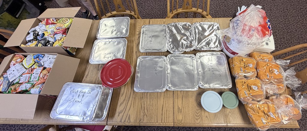 A table holds aluminum foil-covered food trays, plastic bags, and boxes with various items. Some food is on plates.