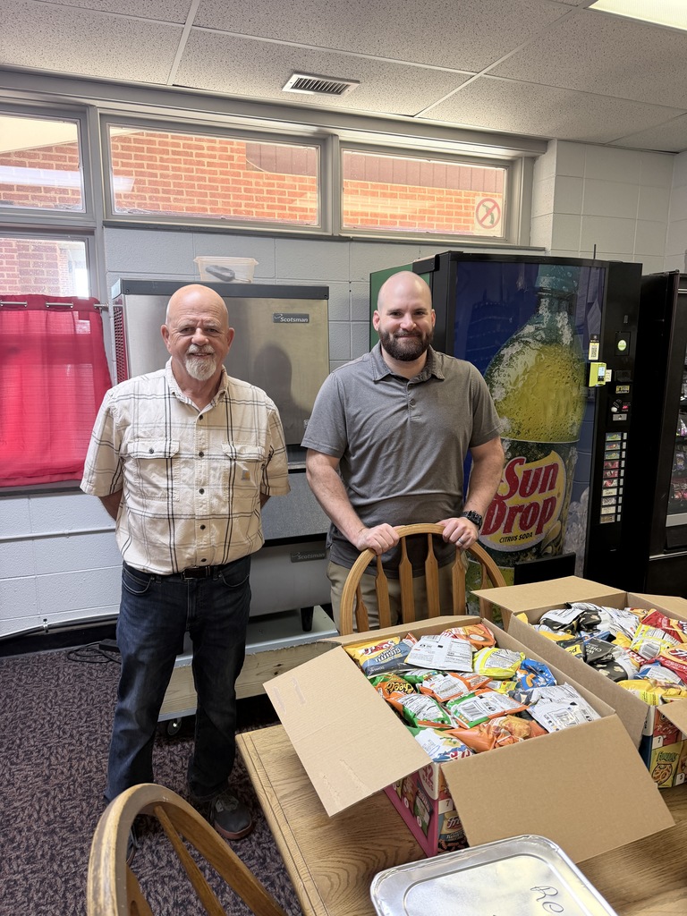 Two men stand next to a table with boxes of food in front of a vending machine.