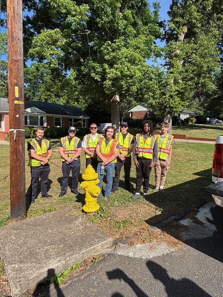 A group of workers in yellow vests, standing next to a fire hydrant, with a grassy area and trees in the background.