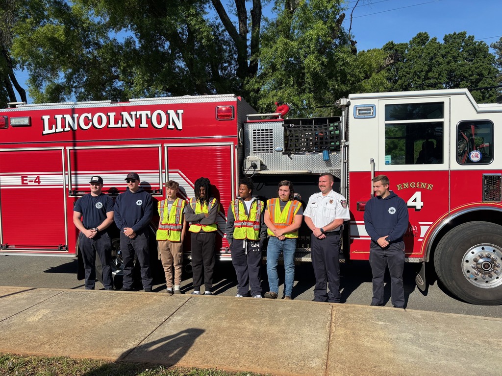Eight people in high-visibility vests stand in front of a fire truck. Trees are visible in the background.