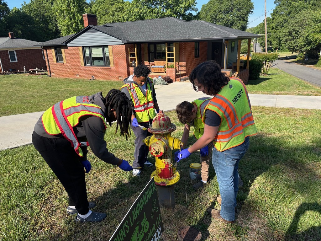 Four people in reflective vests and gloves work on a yellow fire hydrant in front of a house.