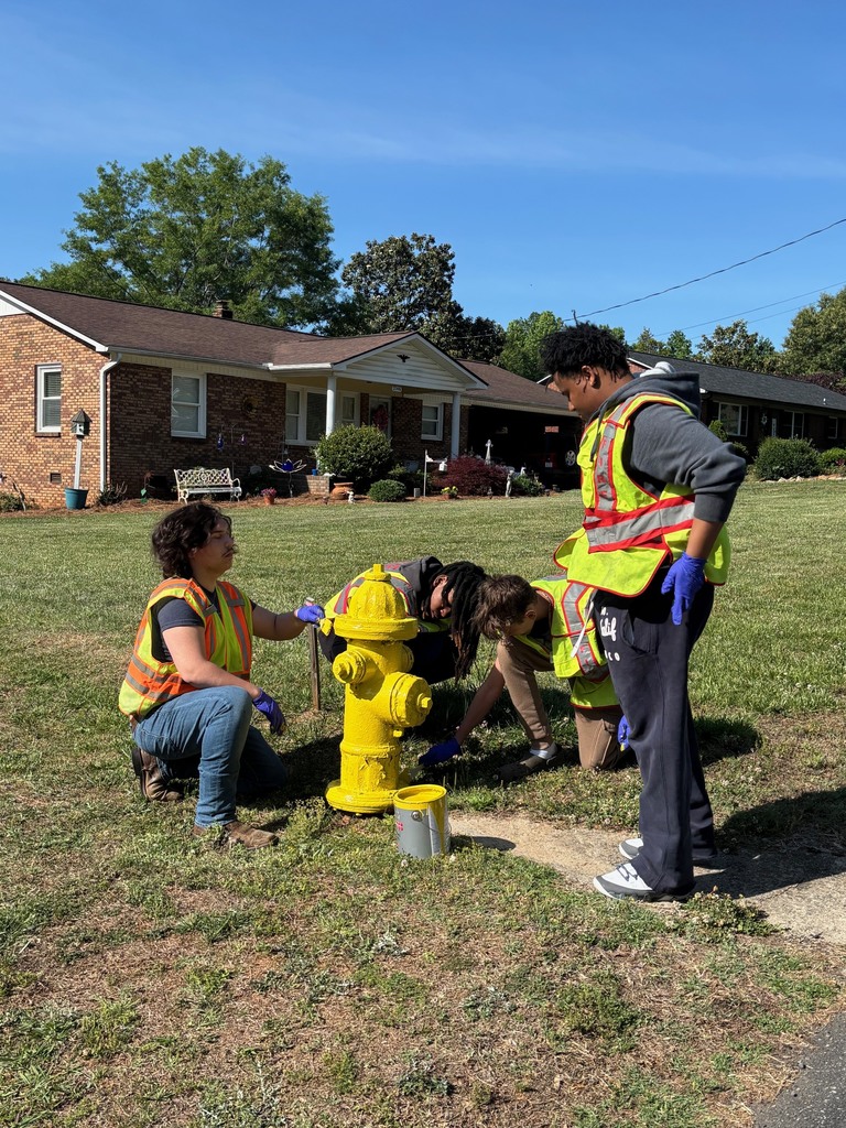 Four people in safety vests and gloves work on a yellow fire hydrant on a lawn.