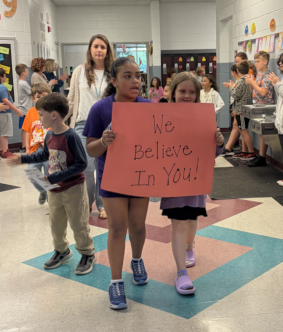 Children in a school hallway hold a sign that says "We Believe In You!" Other students watch.