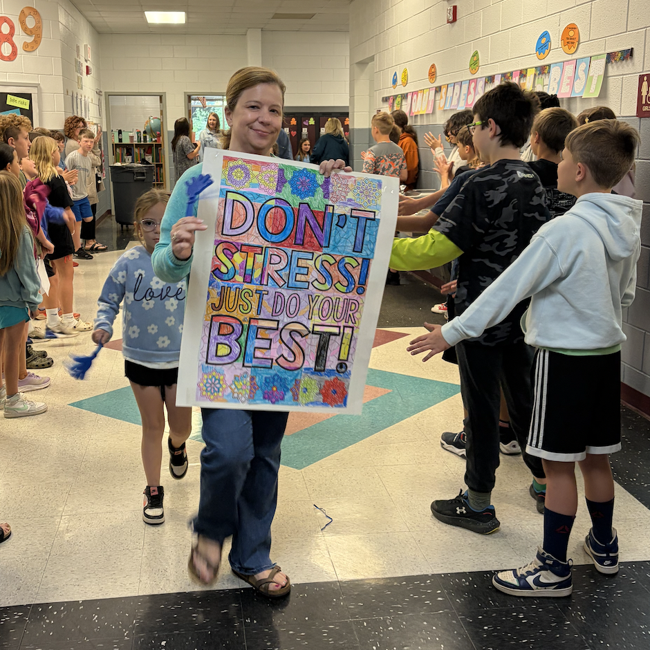 A woman in a hallway holds a colorful sign with the text "Don't Stress! Just Do Your Best!" Children walk nearby.