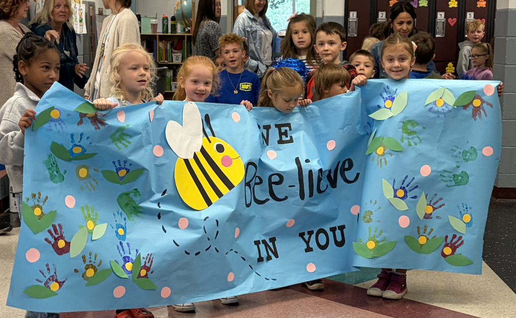 A group of children hold a blue banner decorated with flowers, a bee, and handprints, with "We Bee-live in You" written on it.