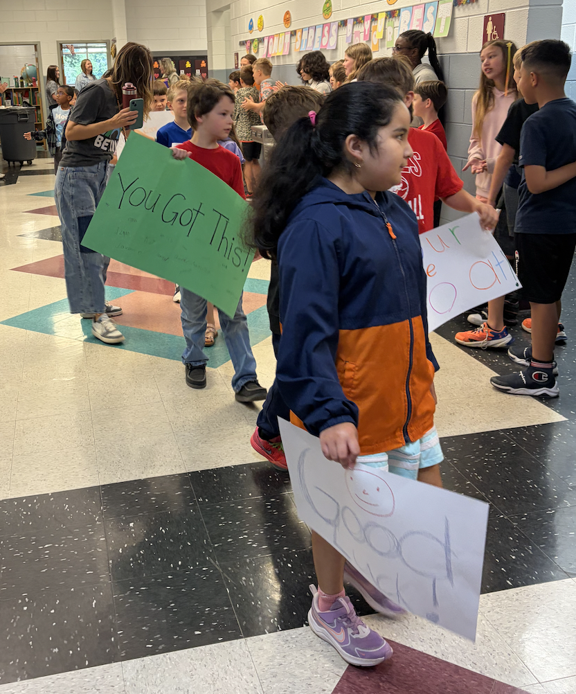 Children walking in a school hallway, holding signs. One child's sign reads "You Got This" and another says "Good Job."