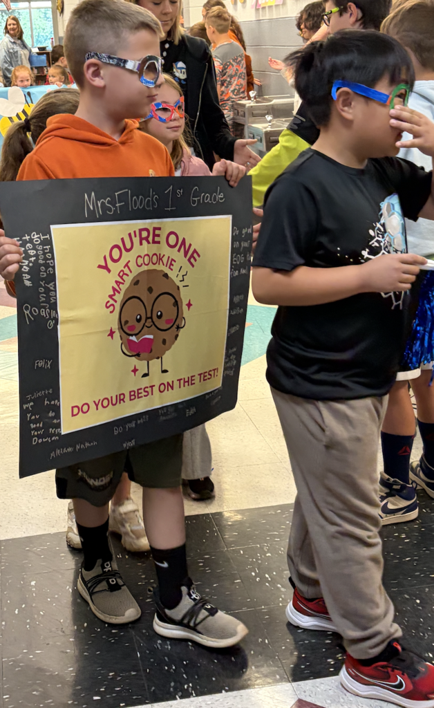 Two boys in a hallway, one holding a sign that reads, "You're a Smart Cookie," while others are in the background.