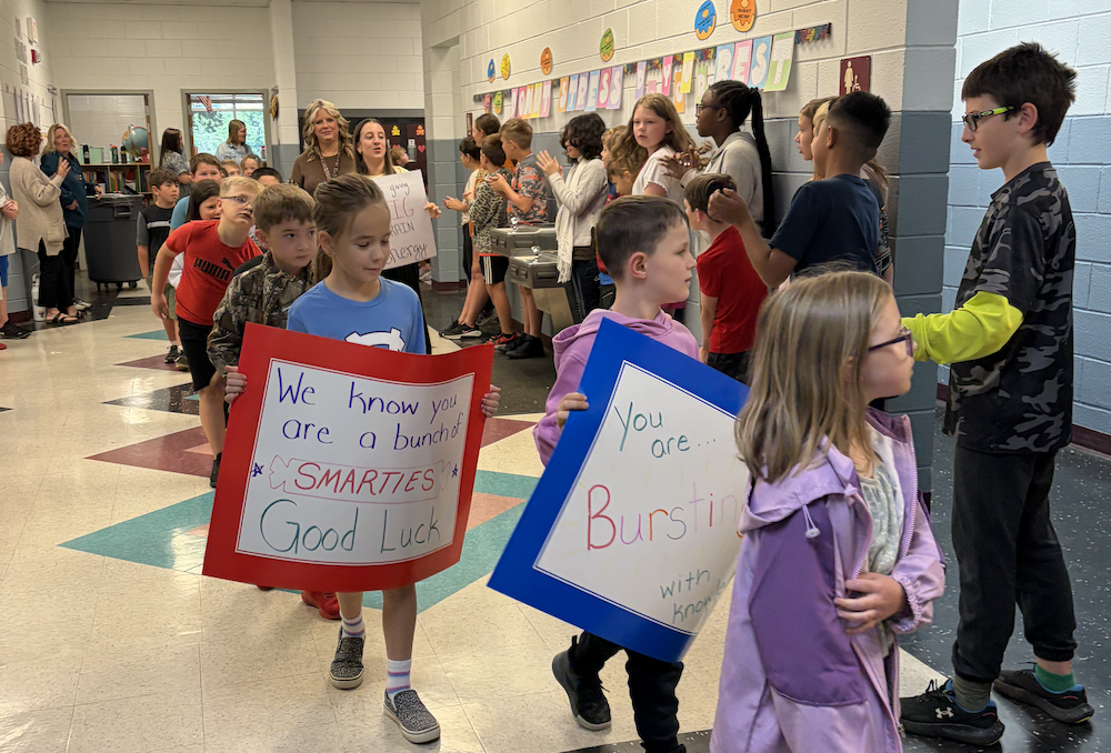 Several children, some holding signs, are walking through a hallway. Some signs read "We know you are a hero".