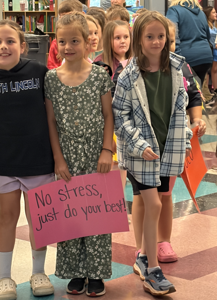 Three young girls standing in a school hallway. One holds a pink sign that says, "No stress, just do your best!"