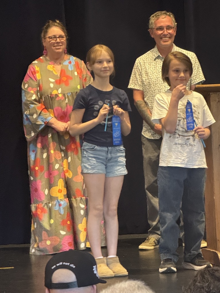 Three children and a woman on stage, holding ribbons. A man stands behind them, and the audience watches.