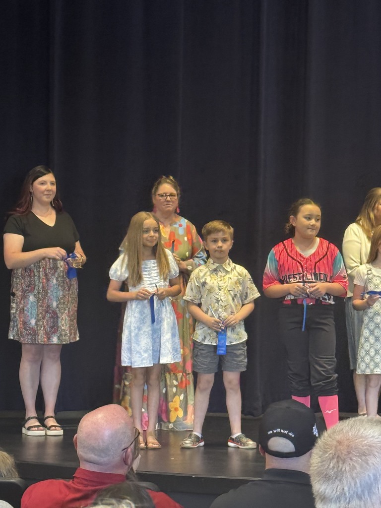 Group of children and adults stand on stage with blue sashes around their necks. Audience sits below.