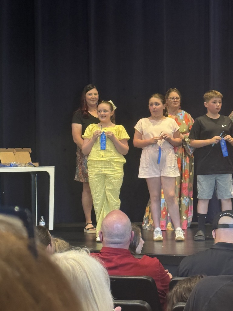 A group of children, some with awards, stand on stage with adults. They are in front of a black curtain.
