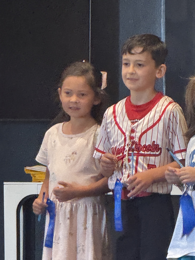 Three children stand in front of a dark background. A girl holds a blue ribbon, a boy holds one too.