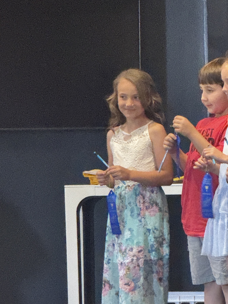 Three children stand together in front of a dark background. They each hold blue ribbons.