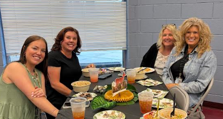 Four women seated at a table, smiling. The table holds dishes, drinks, and a dessert. A window and brick wall in the background.