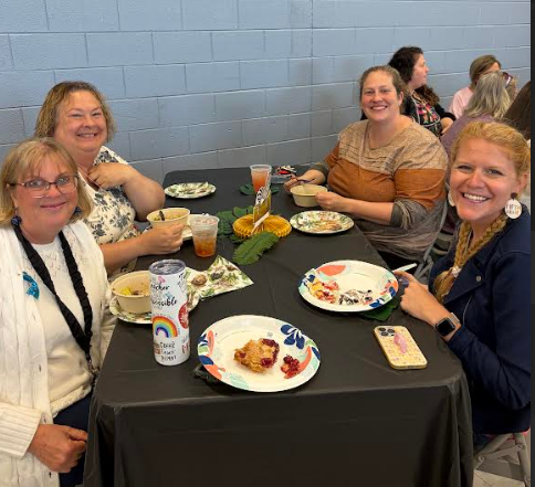 Five women seated at a table, smiling. Food, drinks, and utensils on the table. Wall in background.