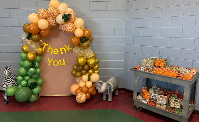 Decorated archway with balloons and the words "Thank You" on it. A table with fruits and a shelf with boxes nearby.