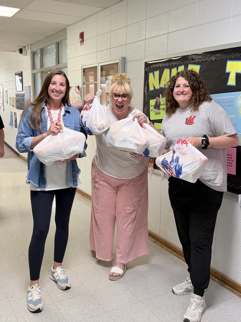 Three women stand in a hallway holding white bags; one holds a blue jacket. Behind them is a bulletin board with text.