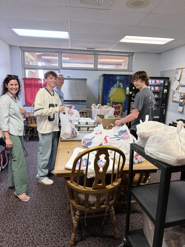Four individuals in a room with tables, chairs, and shelves. Two people hold white bags, and a vending machine is visible.