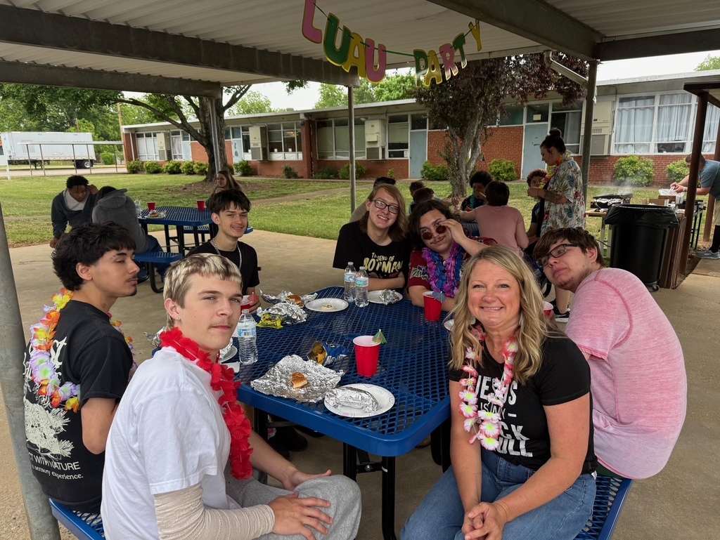 A group of people at a picnic table, some wearing leis, with a building in the background.