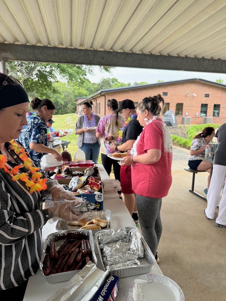 People are gathered at an outdoor buffet table under a shelter, with food, a building, and trees in the background.