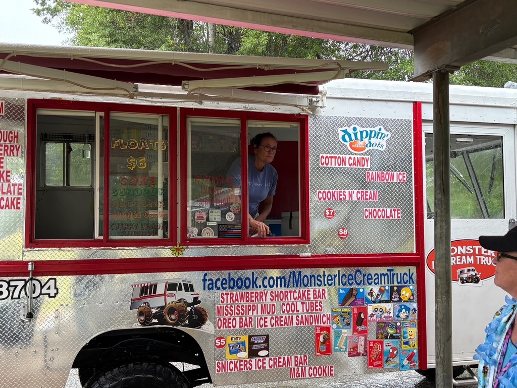 A person in a blue shirt serves ice cream from a window of a white ice cream truck with multiple stickers.