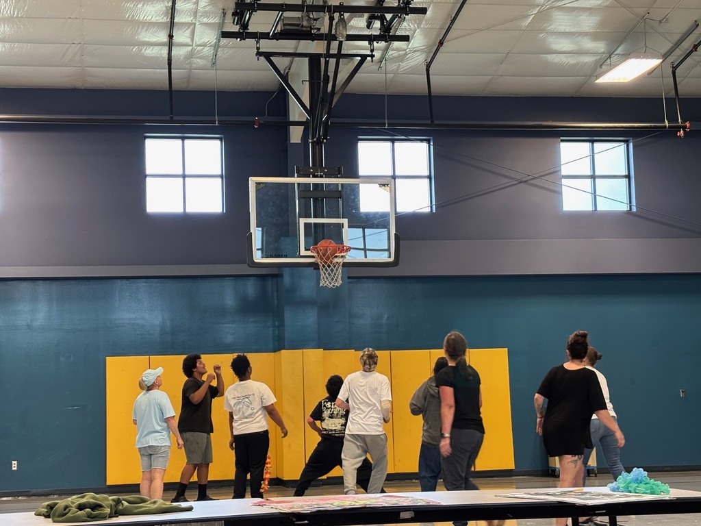 People in a gymnasium with a basketball hoop. A group stands in a circle, and others are near the table.