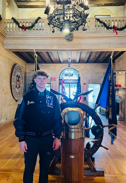 A person stands in front of a brass helm with a flag. Behind them, there is a wooden floor, and a chandelier.