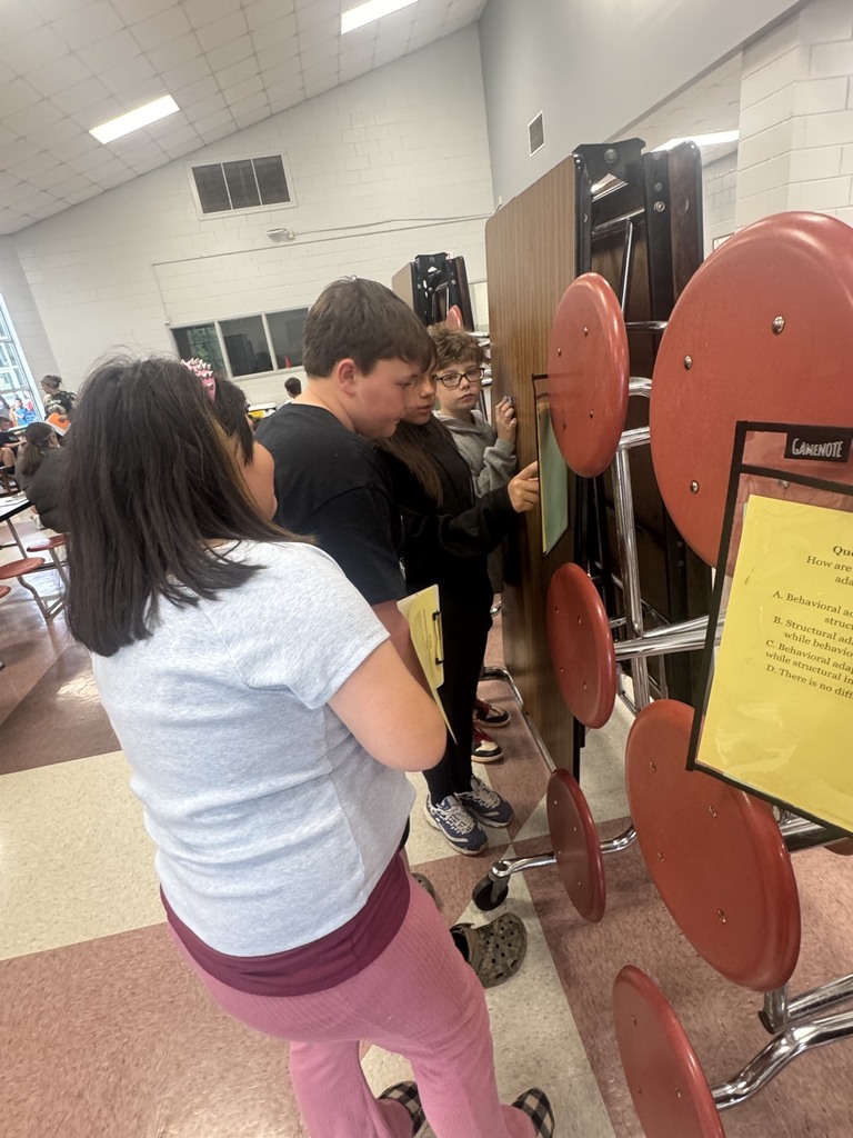 Several students examine a bulletin board with red chairs behind them. A yellow sign is in front of the board.