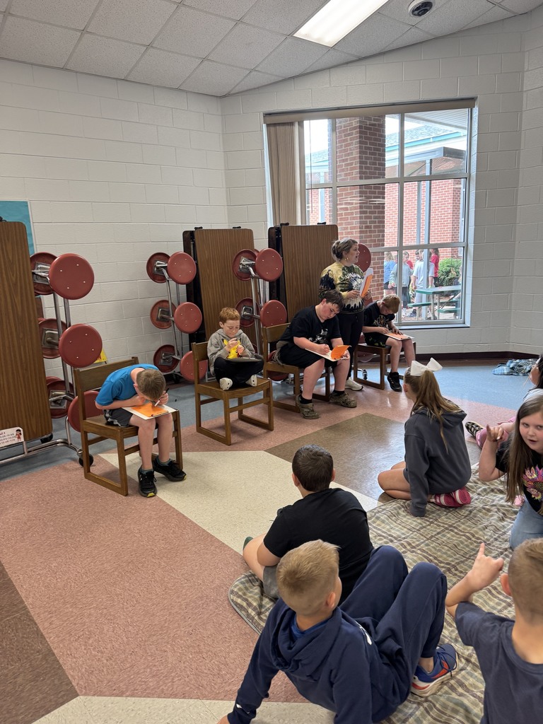 A group of children sits around a room on a carpet, engaging in activities with books and snacks.