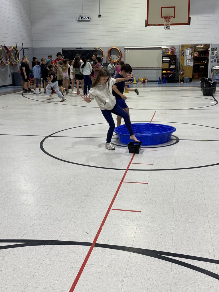 A group of children are playing in a gymnasium with a basketball hoop, a blue water container, and red and black markings on the floor.