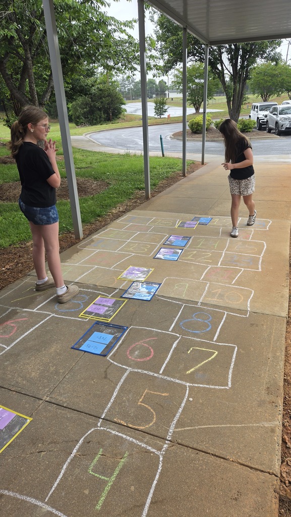 Two individuals play hopscotch on a sidewalk, surrounded by trees and parked cars, with numbered squares and a car-filled parking lot in the background.
