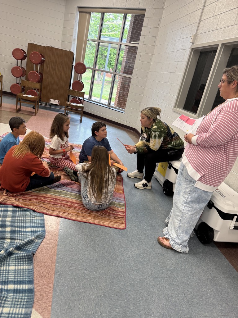 A group of people, including adults and children, sit in a circle on a rug. An adult stands nearby.