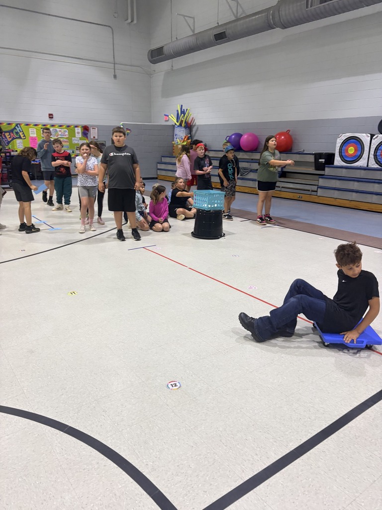 Multiple individuals are in a gymnasium with some seated or standing and others engaging in activities.