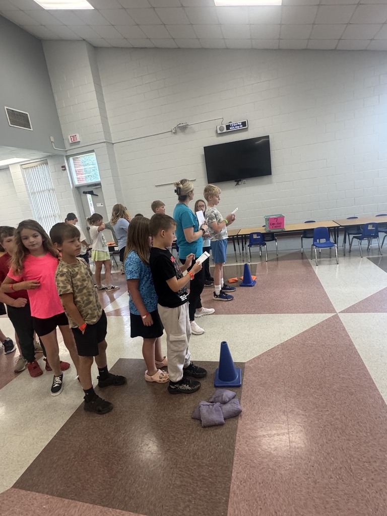 A group of children stands in a hallway. A few hold books and some wear sneakers. Cones are on the floor.