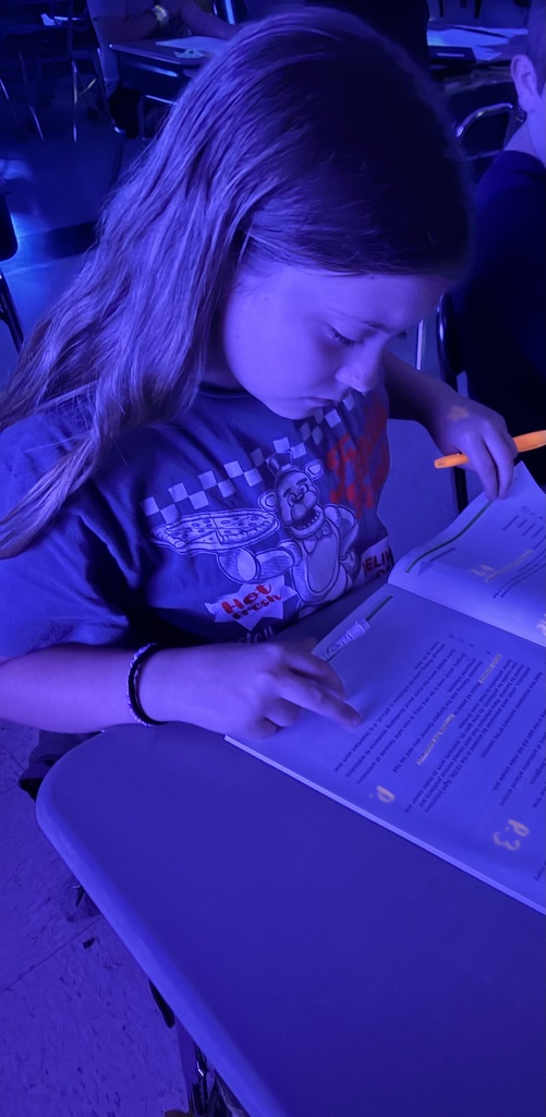 A young girl, wearing a t-shirt, reads a book while seated at a table. She holds an orange marker.