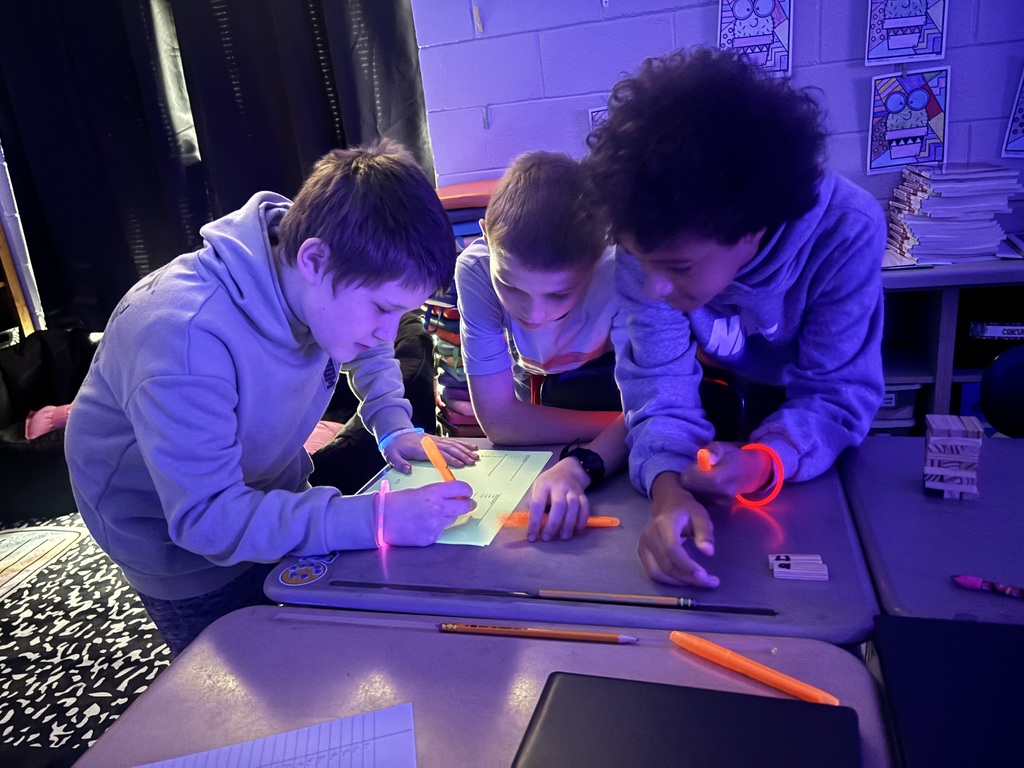 Three young people in a classroom with black curtains behind them are looking at papers and writing.