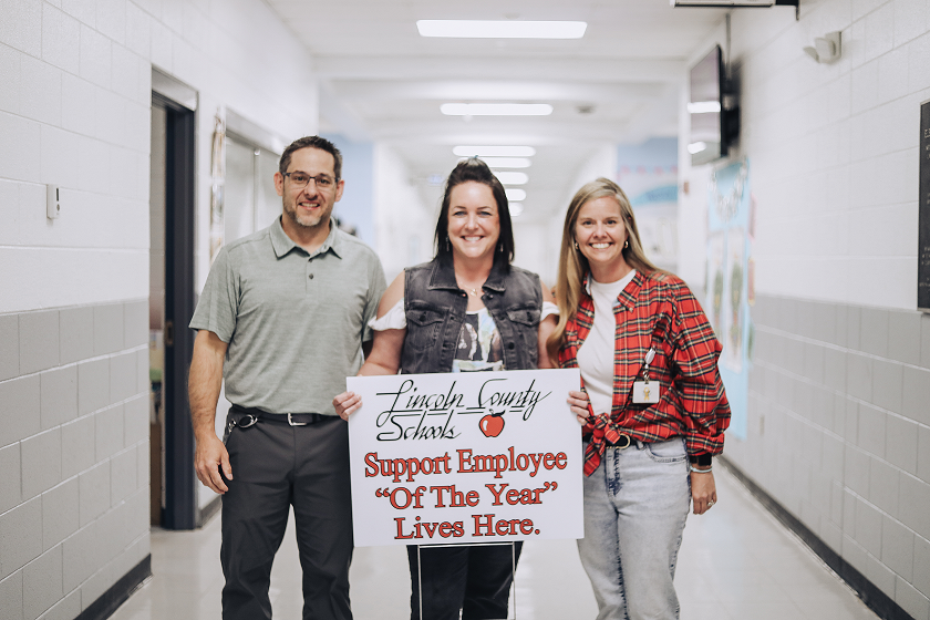 Three smiling Lincoln County Schools staff members stand in a school hallway. The man on the left wears a grey polo shirt and dark trousers. The woman in the center holds a white sign that reads, Lincoln County Schools Support Employee "Of The Year" Lives Here, featuring a small red apple icon. She wears a denim vest over a white top. The woman on the right wears a red and black plaid flannel shirt and light-colored jeans. The hallway behind them has light-colored brick walls and recessed ceiling lights.