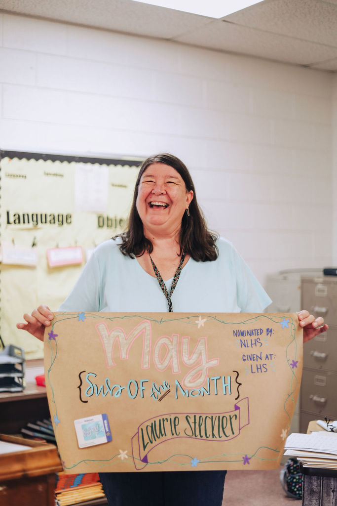 Woman holding a cardboard sign that says "May" with a smiling face in a classroom.