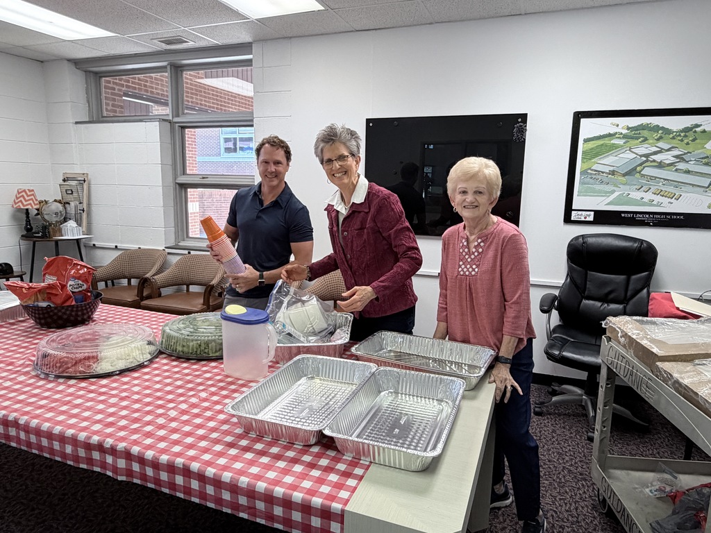 Three people prepare food in a room. A table holds containers and foil pans. A television and picture are on the wall.
