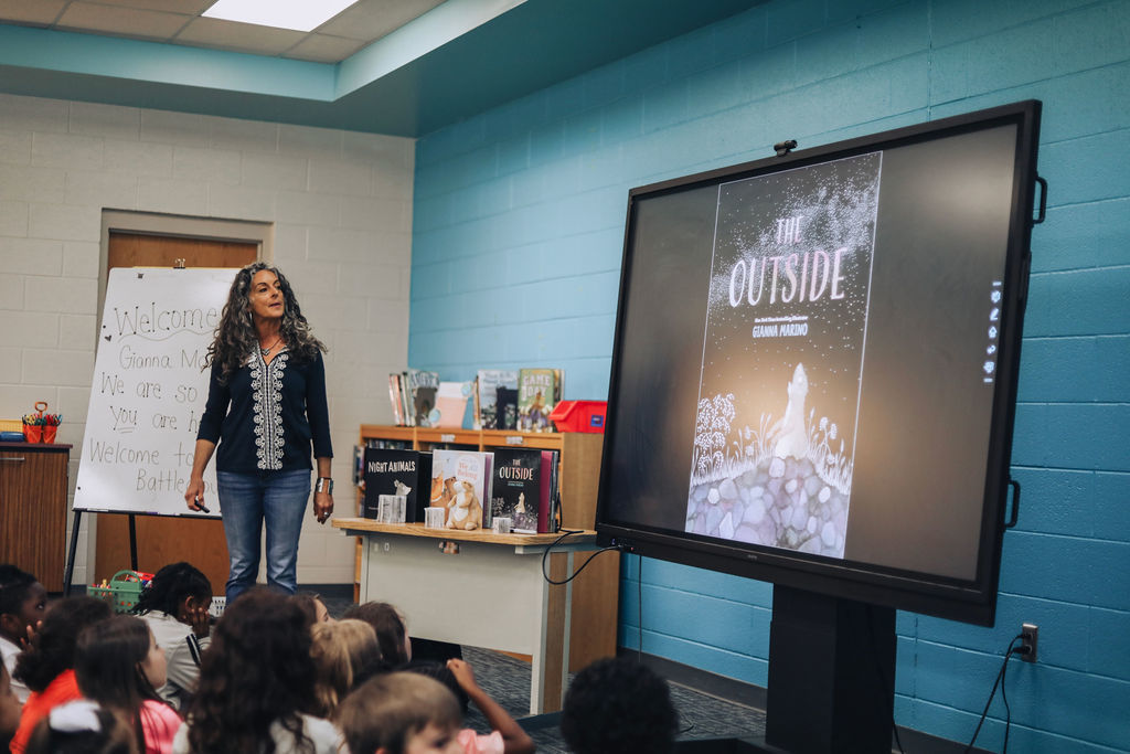 Woman stands in front of classroom, presenting to children seated on floor; monitor displays book cover.