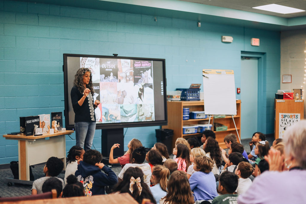 A woman stands before a screen, addressing a room of children seated on the floor.