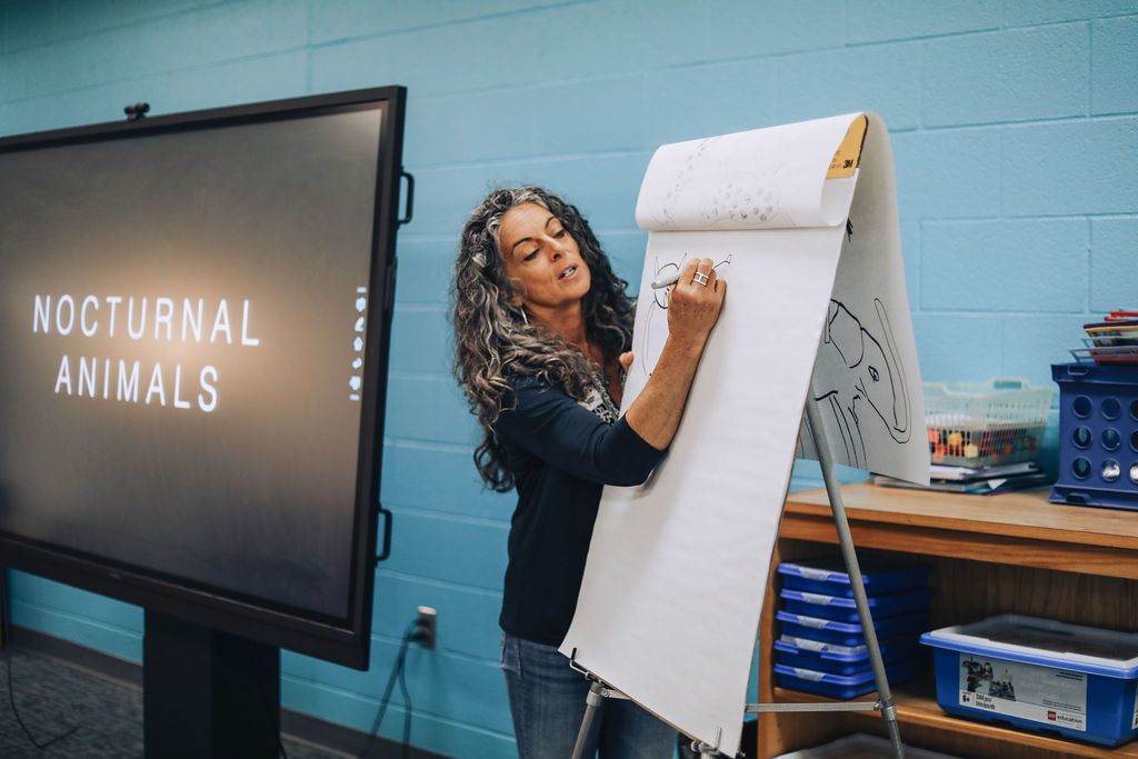 Person standing by a flip chart, writing. A monitor displays text reading "Nocturnal Animals." A desk and blue bins are behind.