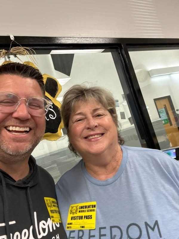 Close-up photo of a man with glasses and a woman smiling indoors. Both are wearing yellow "Lincolnton Middle School Visitor Pass" stickers on their shirts.