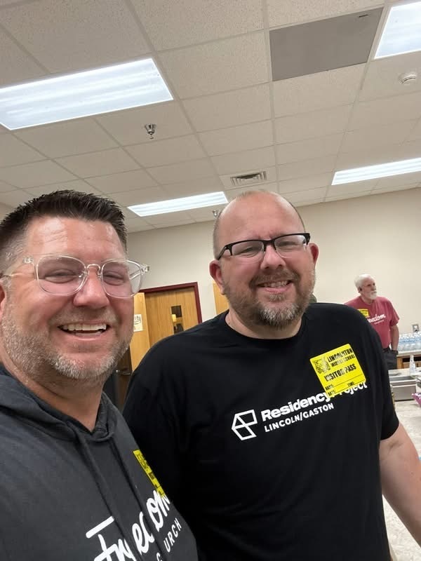 A man with glasses and a man with a beard and glasses smiling for a photo. Both are wearing yellow Lincolnton Middle School visitor passes. The man on the right is wearing a black shirt that says "Residency Project Lincoln/Gaston."