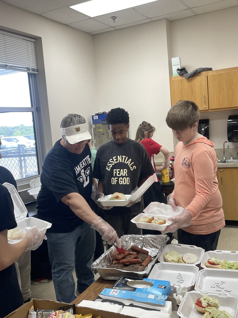 An adult and two students standing at a table preparing food. The adult is wearing a white visor and a "Team Beattie" shirt, using tongs to serve hot dogs and burgers from a foil tray into foam containers.